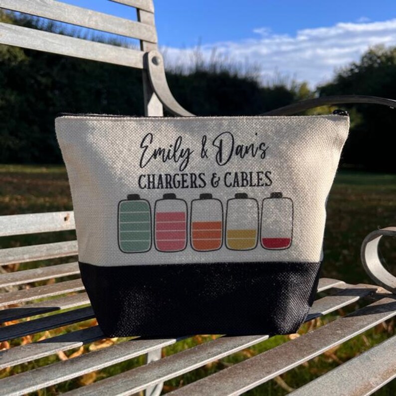 May include: A white canvas zippered pouch with black trim. The pouch has the text "Emily & Dan's Chargers & Cables" printed on the front. Five colorful battery icons are printed below the text.