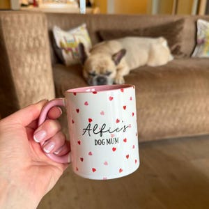 May include: White ceramic mug with a pink handle and interior, decorated with red and pink hearts. The mug has the text "Alfie's DOG MUM" in black script. A dog is resting on a brown sofa in the background.
