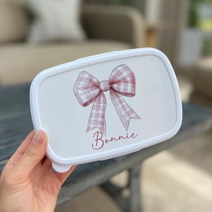 May include: White rectangular lunchbox featuring a pink gingham bow and the name "Bonnie". The lunchbox is held by a hand. The background is blurred, with a wooden table in the foreground.