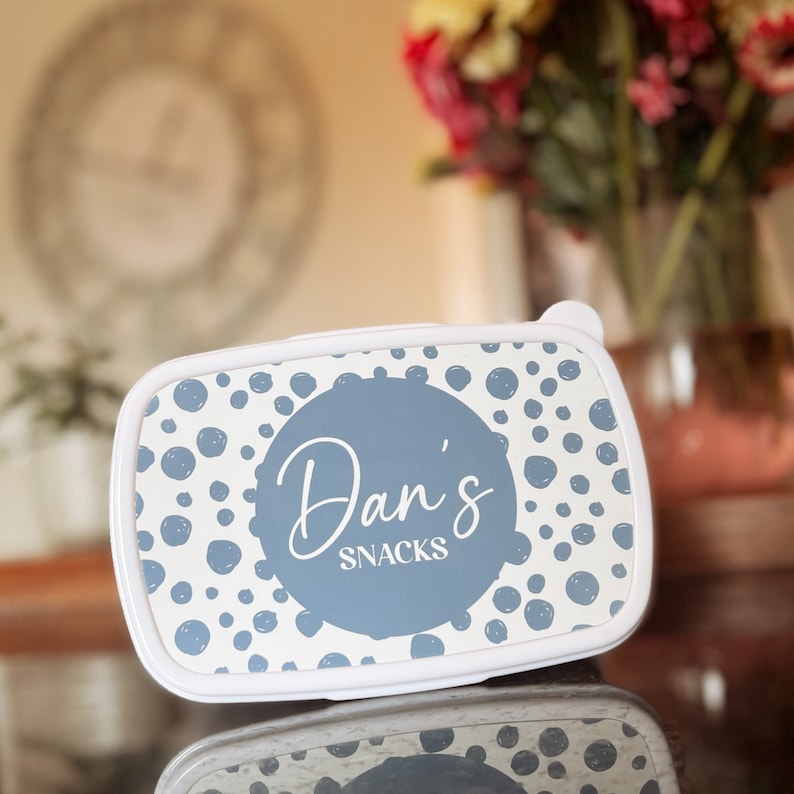 May include: White lunchbox with a blue and white polka dot pattern. The lid has a large blue circle with the text "Dan's Snacks" in white.