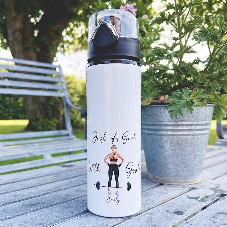 Op de afbeelding: Een witte waterfles van roestvrij staal met een zwarte dop en een ontwerp met de tekst "Just a girl with goals" met een vrouw die gewichten heft en de naam "Emily" onderaan.