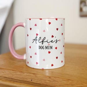 May include: White ceramic mug with a pink handle and interior, adorned with scattered red and pink hearts. The mug features the text "Alfie's DOG MUM" in black script, making it a charming gift for dog lovers.