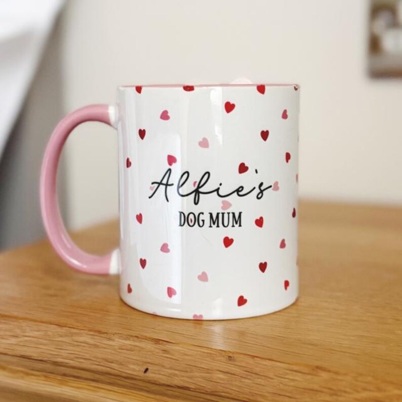 May include: White ceramic mug with a pink handle and interior, adorned with scattered red and pink hearts. The mug features the text "Alfie's DOG MUM" in black script, making it a charming gift for dog lovers.