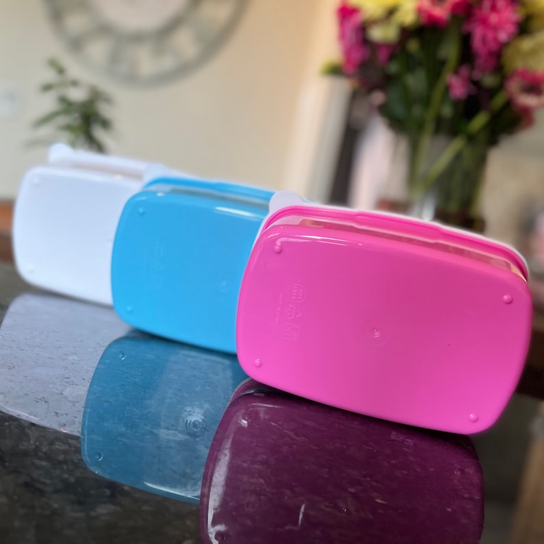 May include: Three plastic lunch boxes, one white, one blue, and one pink. The boxes are stacked on top of each other.