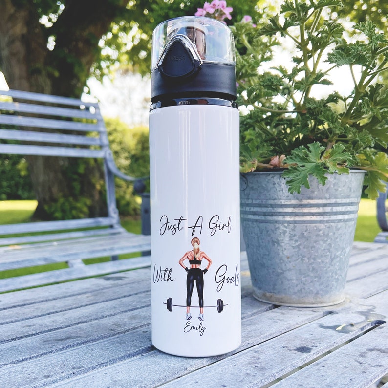Op de afbeelding: Een witte waterfles met een zwarte dop en een zwart-witte illustratie van een vrouw die gewichten heft. De tekst "Just A Girl With Goals" staat op de fles, samen met de naam "Emily".
