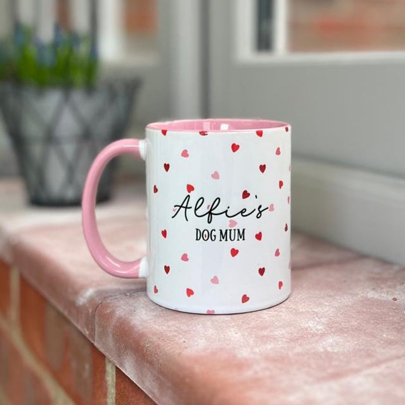May include: White ceramic mug with a pink handle and interior, decorated with red and pink hearts. The mug features the text "Alfie's DOG MUM" in black script. A charming gift for dog lovers.