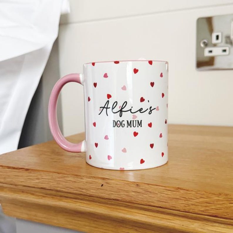 May include: White ceramic mug with a pink handle, adorned with red and pink heart patterns. The mug features the text "Alfie's DOG MUM" in a script font. The mug is placed on a wooden surface.