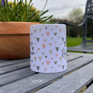 Puede incluir: Taza de cer&aacute;mica blanca con un patr&oacute;n de corazones rosas y grises. La taza tiene el texto "A Dog's Mum" en el lado derecho y "Love is" en el lado izquierdo.
