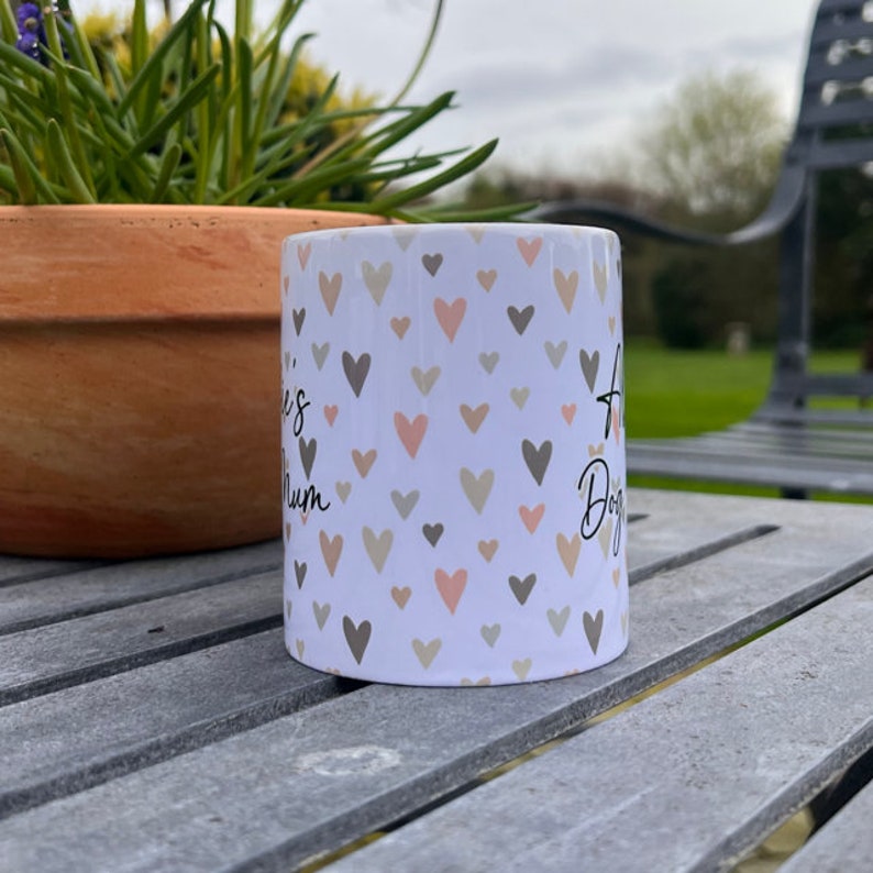 Puede incluir: Taza de cer&aacute;mica blanca con un patr&oacute;n de corazones rosas y grises. La taza tiene el texto "A Dog's Mum" en el lado derecho y "Love is" en el lado izquierdo.