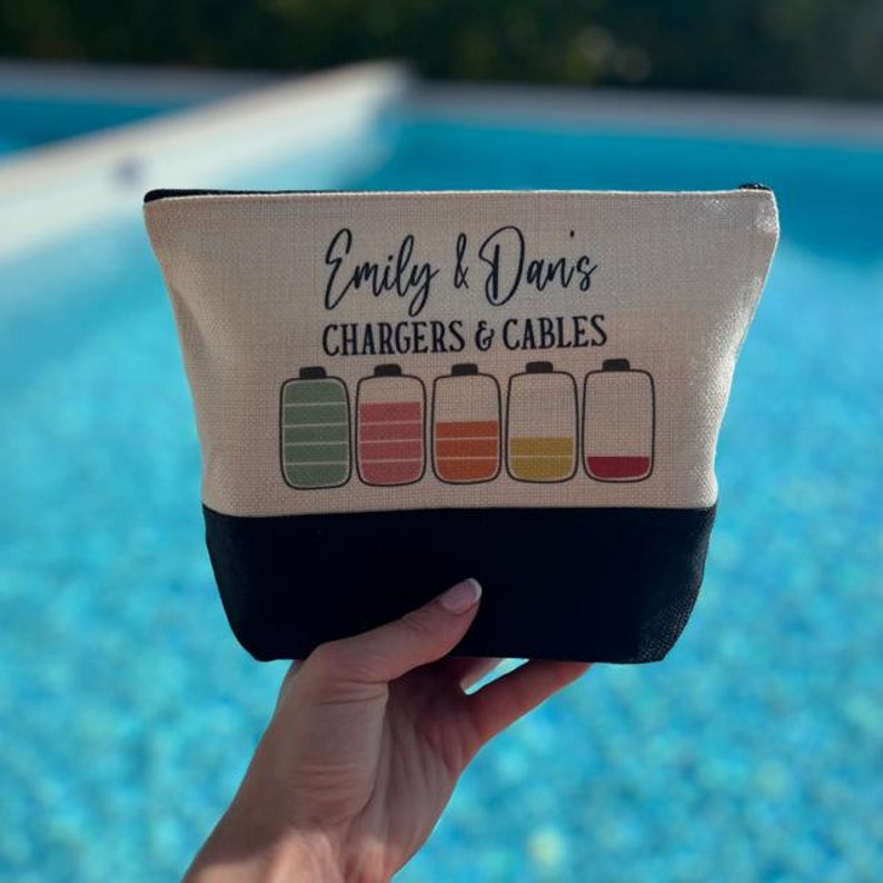 May include: A beige canvas makeup bag with black trim. The bag has the text "Emily & Dan's Chargers & Cables" printed on it. Five colorful battery icons are below the text.