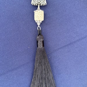 May include: A silver-toned pendant with a cream-colored, cylindrical bead and a long black tassel. The pendant has an ornate top piece with a decorative design. The tassel is made of many strands of black thread.