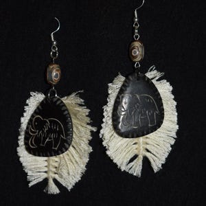May include: A pair of handcrafted earrings featuring a black, teardrop-shaped pendant with an etched elephant design. The pendants are accented with cream-colored feather-like fringe and a small, patterned bead. The earrings hang from silver-colored hooks.