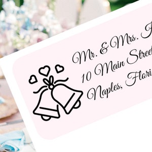 Könnte beinhalten: Ein weißes Hochzeit-Adress-Etikett mit schwarzem Text und einer schwarz-weißen Illustration von zwei Hochzeitsglocken mit Herzen. Der Text lautet "Mr. & Mrs. Henry Knight, 10 Main Street, Naples, Florida 34110."