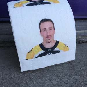 May include: A white toilet paper roll with a printed image of a man wearing a yellow and black hockey jersey. The man is looking directly at the camera.
