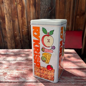 May include: A rectangular white metal storage tin featuring the orange text RYKRISP vertically on the side. The design includes colorful illustrations of a sliced apple, a rolled slice of meat, a piece of Swiss cheese with an olive, and crackers.