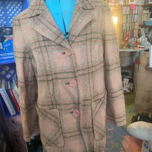 May include: A vintage pink plaid coat with a classic design. The coat features a collared neckline, three pink buttons, and two front pockets. The plaid pattern includes shades of pink, green, and brown. The coat is displayed on a blue dress form.