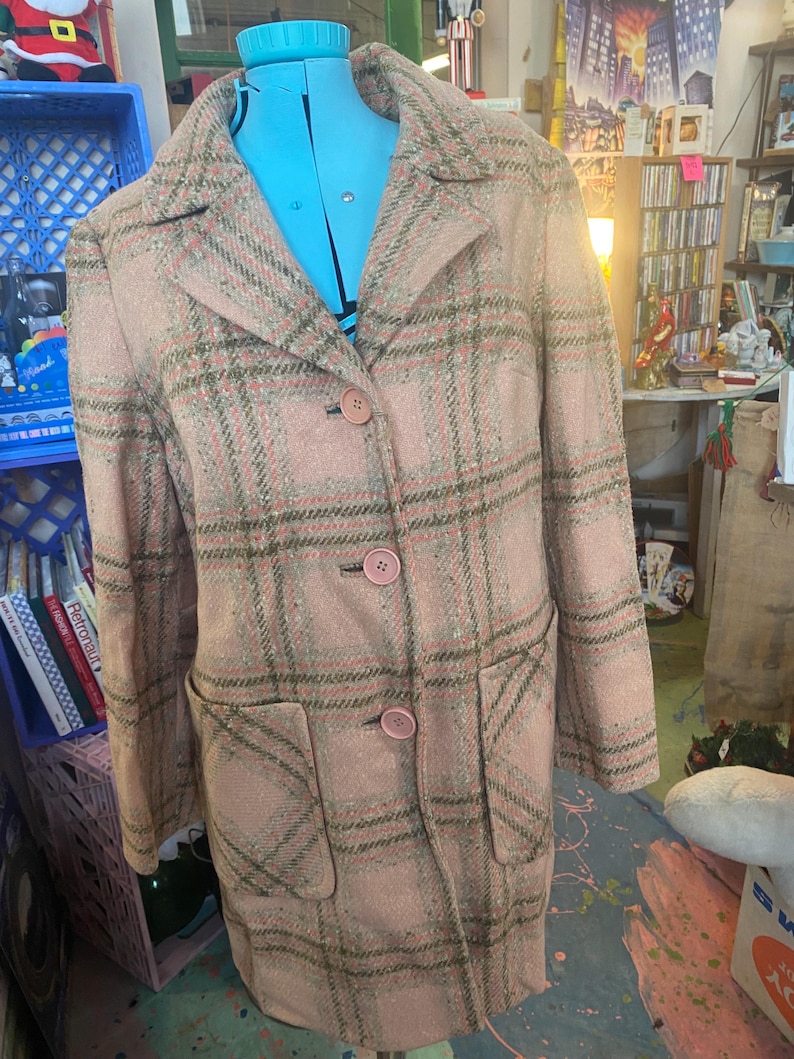 May include: A vintage pink plaid coat with a classic design. The coat features a collared neckline, three pink buttons, and two front pockets. The plaid pattern includes shades of pink, green, and brown. The coat is displayed on a blue dress form.