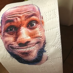 May include: A close-up of a printed toilet paper roll featuring a smiling man's face. The man has a dark complexion and a short beard.