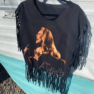 May include: Black t-shirt with a graphic of a singer holding a microphone, with the name "Adele" printed in gold. The shirt has fringe detailing along the sleeves and bottom hem, hanging from a black hanger.