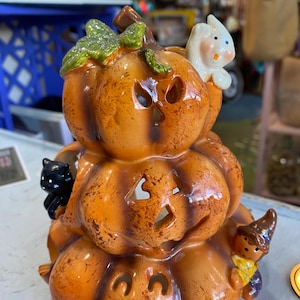Puede incluir: Decoración de Halloween de cerámica de tres niveles con forma de calabazas apiladas. Las calabazas naranjas tienen caras talladas y están adornadas con un gato negro, un fantasma blanco y una pequeña figura con disfraz de bruja.