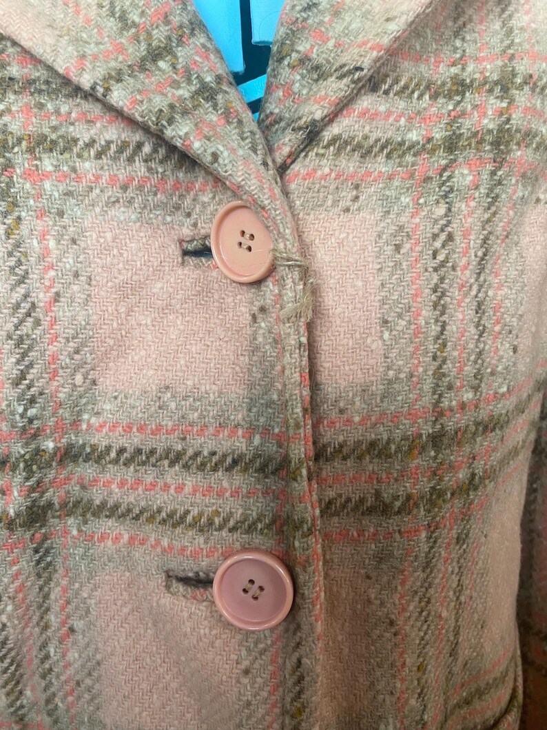 May include: Close-up of a pink and gray plaid jacket with two matching pink buttons. The jacket has a classic collar and a tailored fit. The fabric appears to be a woven wool or wool blend.