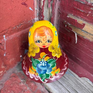 May include: A bell-shaped wooden figurine featuring a hand-painted face with blue eyes and blonde hair under a yellow headscarf. The base is deep red with a large blue flower, green leaves, and white decorative wavy lines along the bottom edge.