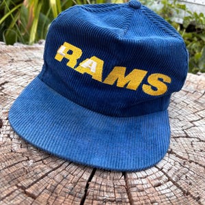 May include: A blue corduroy baseball cap featuring the word RAMS embroidered in yellow with white accents on the front panel. The hat has a structured crown and a curved brim, with a textured fabric finish throughout the entire exterior surface.