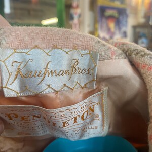 May include: Close-up of a vintage pink and beige wool coat lining. The coat features two tags: one embroidered with "Kaufman Bros." and another with "Pendleton" and "100% Virgin Wool". The coat's interior is a light peach color.