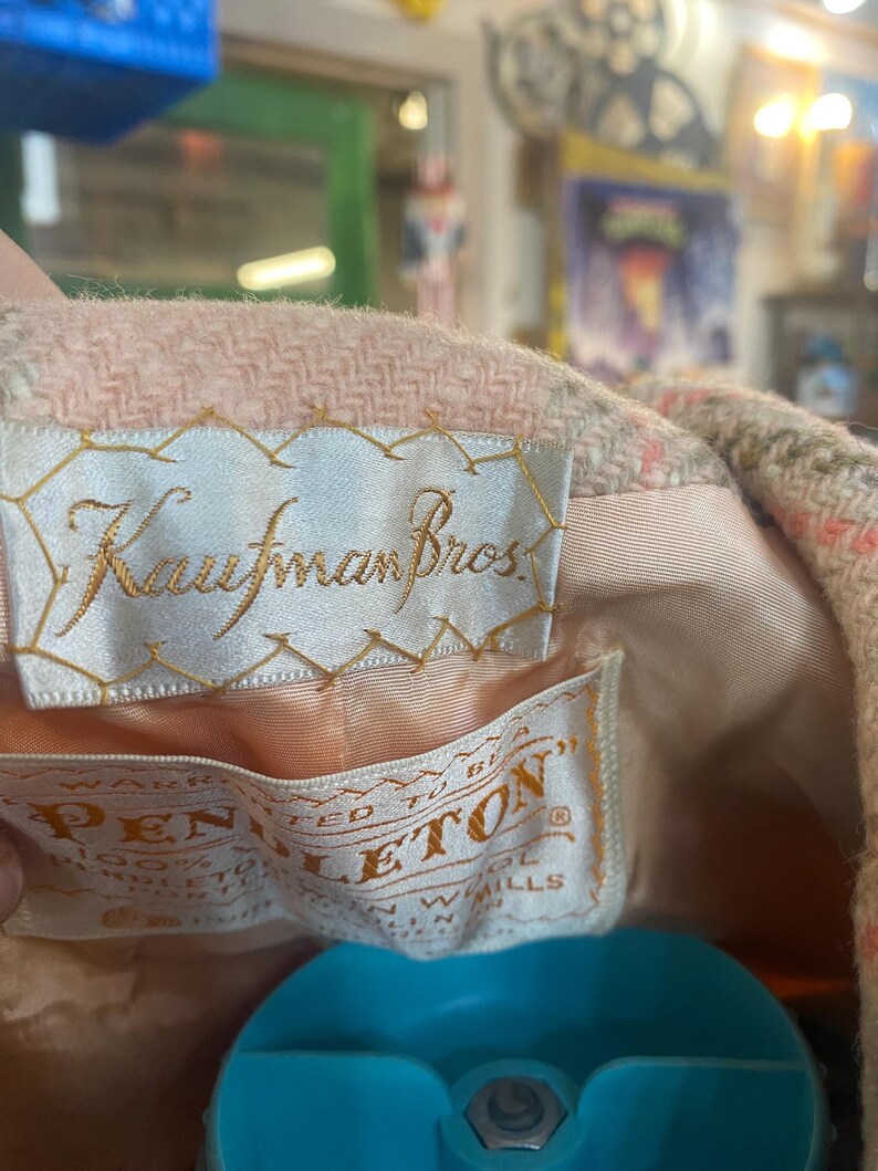 May include: Close-up of a vintage pink and beige wool coat lining. The coat features two tags: one embroidered with "Kaufman Bros." and another with "Pendleton" and "100% Virgin Wool". The coat's interior is a light peach color.