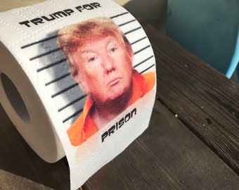 May include: A roll of toilet paper with a mugshot of Donald Trump on it. The text on the toilet paper says "Trump for Prison".