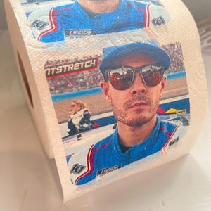 May include: A roll of toilet paper with a printed image of a race car driver wearing sunglasses and a blue racing suit. The driver is wearing a blue and white baseball cap with the word "NASCAR" on it. The background of the image is a race track with a crowd of people in the stands.