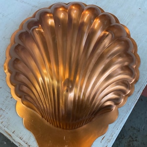 May include: A copper-colored, shell-shaped mold with a scalloped edge. The mold has a detailed, ridged design radiating from the center. The copper has a warm, reflective sheen, suggesting it is made of metal. The mold is likely for baking or decorative purposes.