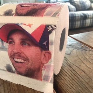 Peut inclure: Un rouleau de papier toilette avec une image imprimée d'un homme portant une casquette de baseball rouge, blanche et bleue. L'homme sourit et regarde vers la droite. Le papier toilette est sur une surface en bois.