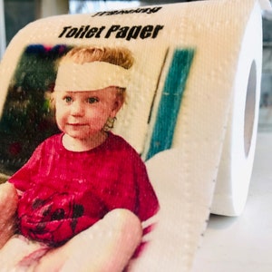 May include: A roll of white toilet paper with a printed image of a young child wearing a red shirt and a white bandage on their forehead. The text "Toilet Paper" is printed on the roll.