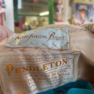 May include: Close-up of two vintage fabric labels. The top label reads "Kaufman Bros." in elegant script. The bottom label says "Pendleton" with "100% Virgin Wool" and "Pendleton Woolen Mills, Portland, Oregon" printed on it.