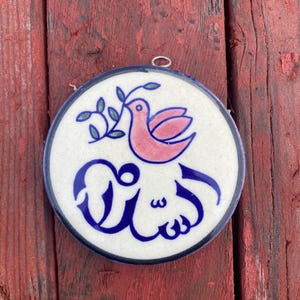 May include: Round ceramic wall hanging with a pink bird holding a green sprig. The bird is above blue Arabic script on a white background, framed by a blue border. A metal loop is at the top for hanging.