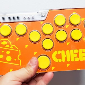 May include: Orange arcade button panel with yellow buttons and the word "CHEESE" printed in yellow. The panel features a yellow cheese graphic.