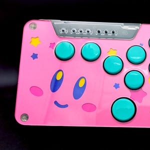 May include: A pink arcade stick with a Kirby character design. The stick has teal buttons and a yellow star design.