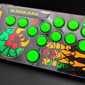 May include: A black and green arcade-style controller with the word "METROID" printed on the side. The controller has 10 green buttons and a black and yellow graphic of a creature from the Metroid video game series.