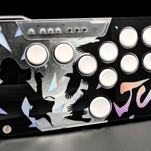 May include: A black arcade controller with white buttons and a holographic design featuring a silhouette of a person and birds. The controller has the word "JUN" written on it in a rainbow font.
