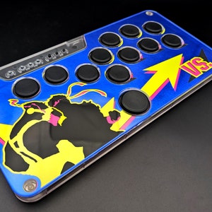 May include: A blue arcade stick with black buttons and a clear top section. The stick features a graphic of a fighting game character with yellow and pink accents, and the word "VS." in pink. The design is on a black background.