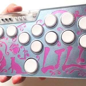 May include: A silver arcade controller with pink floral and cat designs. The controller has 8 white buttons and a clear plastic top with black buttons.