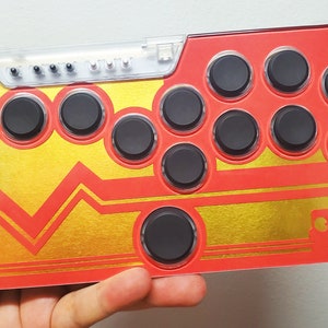 May include: A red and gold arcade controller with 10 black buttons. The controller has a clear plastic top with a row of LEDs.