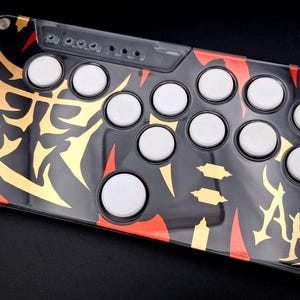 May include: Black arcade game controller with white buttons and a gold and red decorative design. The controller has a clear top layer and is rectangular with rounded corners. The design includes stylized lettering.