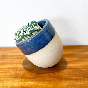 May include: A white and blue ceramic match holder with a rounded shape, filled with green and white matches.