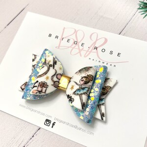 May include: A blue and white glitter hair bow with a ballerina and presents design. The bow is attached to a gold clip.