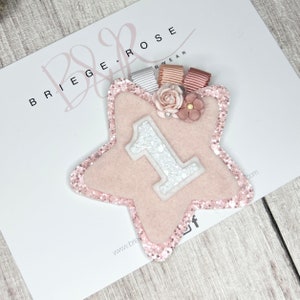 May include: Pink felt star with a white glitter number one on it. The star has a pink glitter border and is decorated with pink and white flowers. The star is on a white background with the text "Briege Rose" in black.