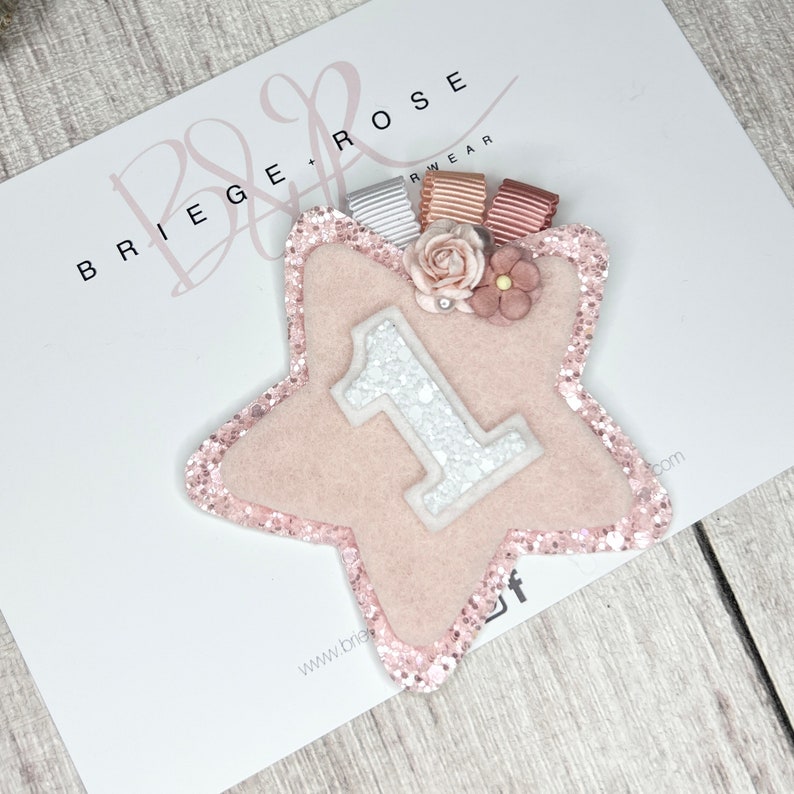 May include: Pink felt star with a white glitter number one on it. The star has a pink glitter border and is decorated with pink and white flowers. The star is on a white background with the text "Briege Rose" in black.