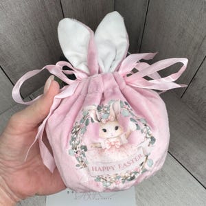 May include: Pink velvet Easter bunny gift bag with white bunny ears and a ribbon. The bag has a printed design of a bunny in a wreath with the text "Happy Easter".