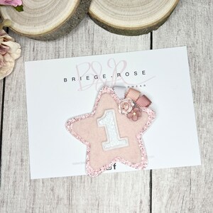 May include: Pink glitter star with a white felt number one and pink and white ribbon embellishments. The star is on a white card with the text "BRIEGE & ROSE WEAR" printed on it.